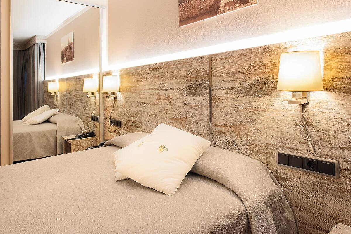 Une chambre d’hôtel avec un grand lit, une tête de lit dotée de lampes fixées au mur, une table de chevet et un miroir qui reflète la pièce.