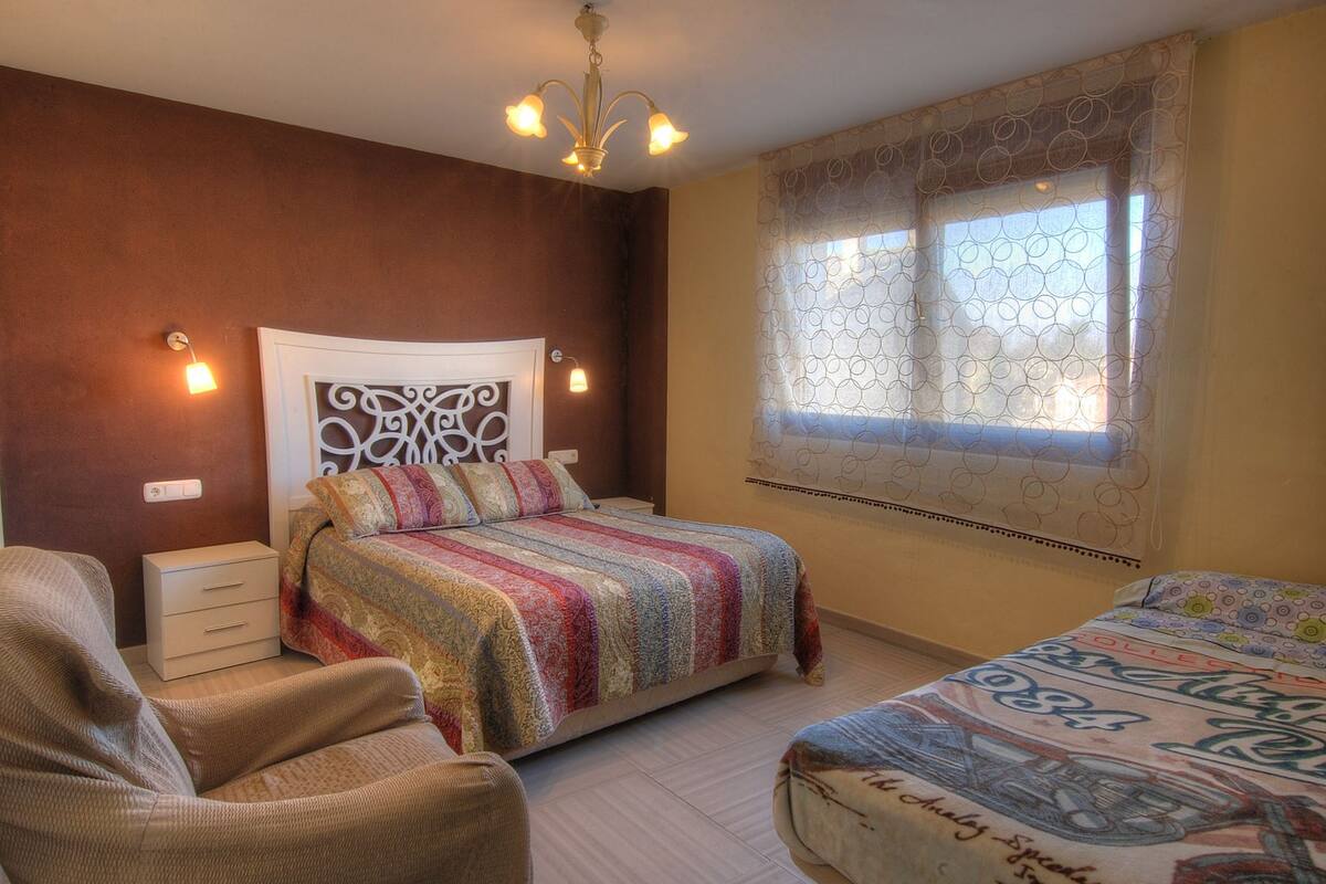 A bedroom with a bed, a chair, a bedside table, a decorative headboard, a window with patterned curtains, and a chandelier.