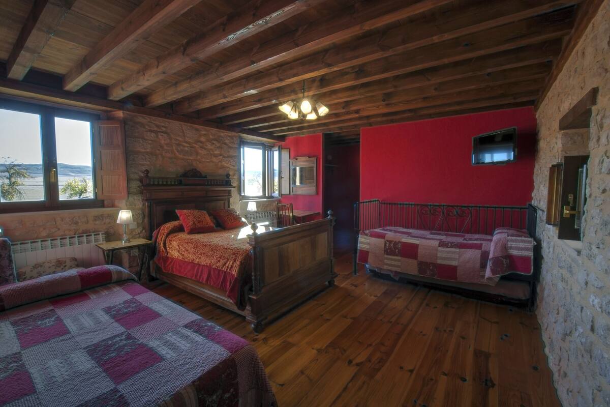 A cozy room with a wooden ceiling, a large bed with a red bedspread, a smaller bed with a quilt, a stone wall, and a window with a view.