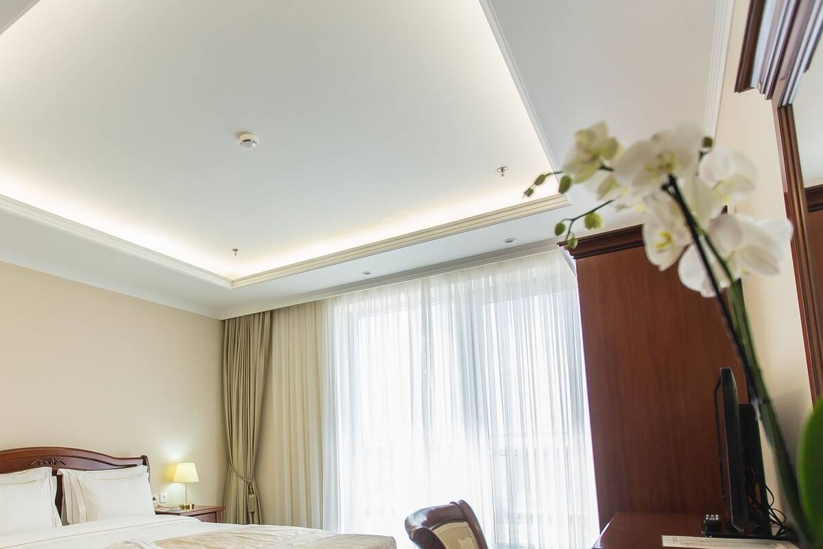 A hotel room with a bed, a desk, a chair, a lamp, and a large window with curtains.
