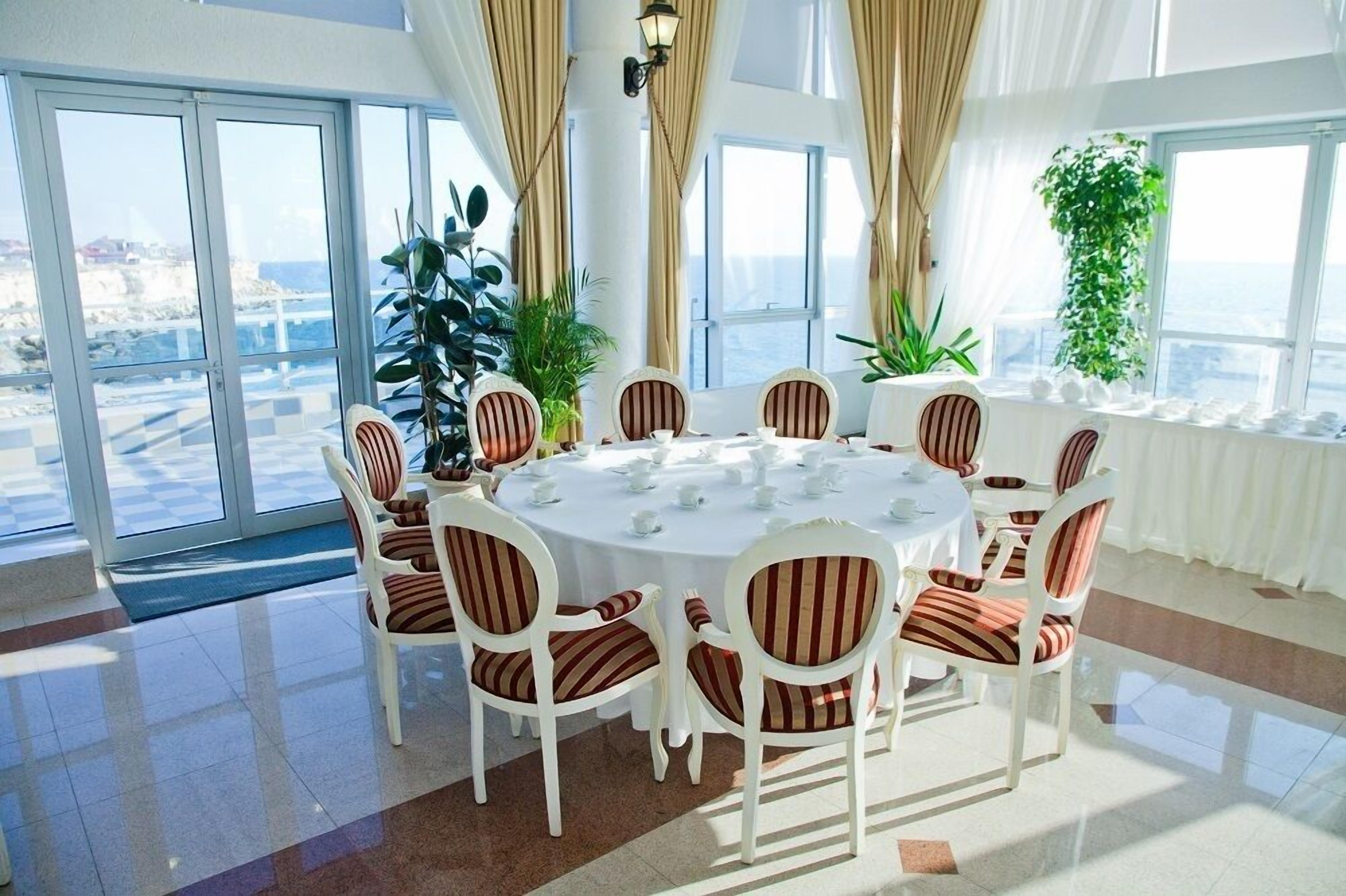 Una mesa redonda para comer con sillas blancas y cojines a rayas, rodeada de ventanas amplias con cortinas, que ofrecen vista al mar.