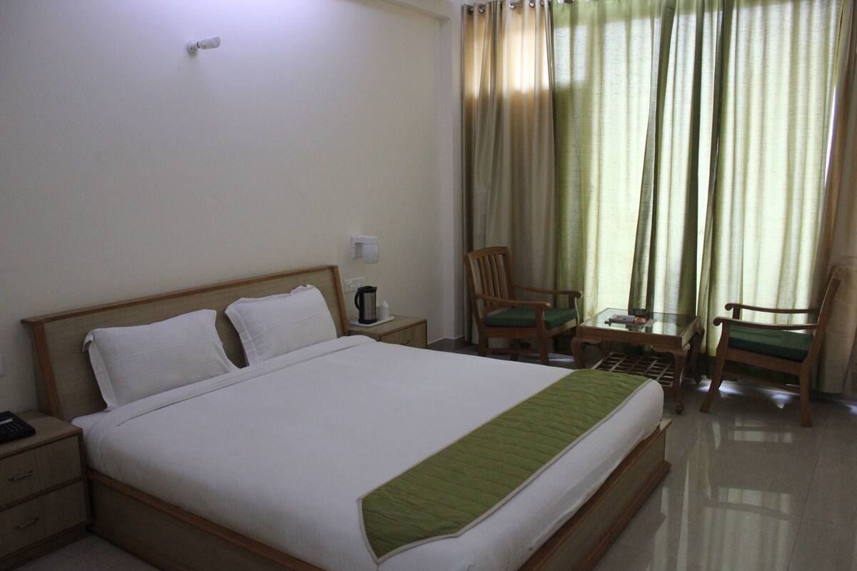 Quarto de hotel com uma cama grande, uma mesa de cabeceira, uma mesa com cadeiras e vista para o exterior através de cortinas.