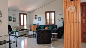A spacious living room with a sofa, armchairs, and a coffee table. There are two windows with wooden frames, a wooden cabinet, and a wall-mounted light fixture. - Magma Guest House (Ragalna)