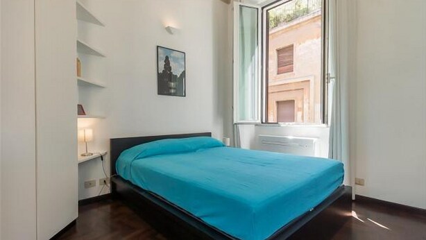Un dormitorio con cama, ventana y estante en la pared con libros.