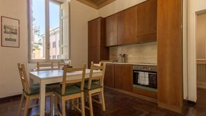 Cocina pequeña con armarios de madera, una mesa de comedor con sillas y una ventana que da a edificios.