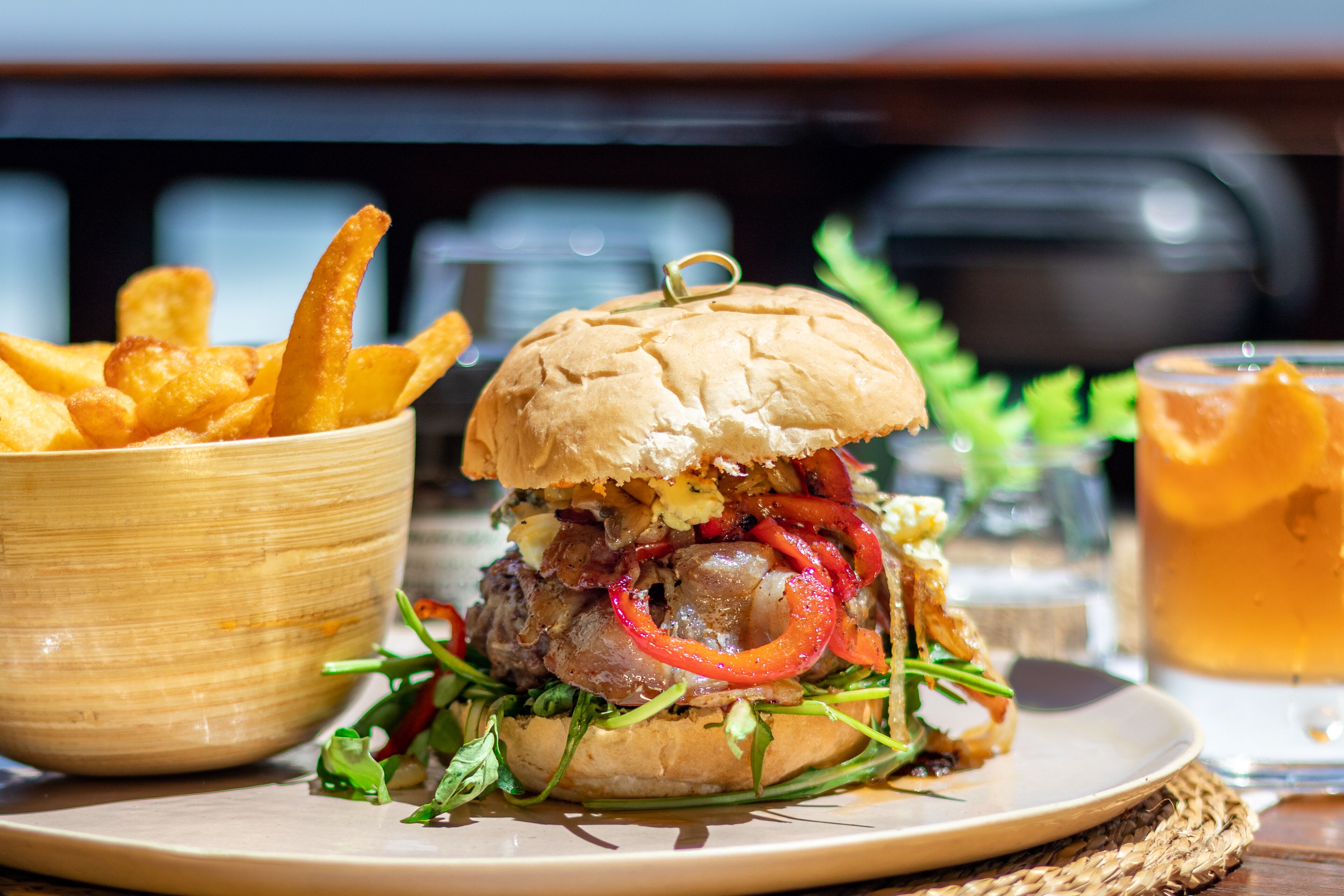 A burger with a bun, lettuce, and red peppers, accompanied by a bowl of fries and a glass of beer on a table.