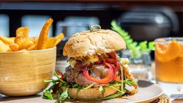 A burger with a bun, lettuce, and red peppers, accompanied by a bowl of fries and a glass of beer on a table.