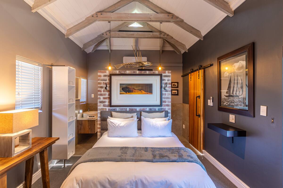 A bedroom with a large bed, a wooden ceiling, a brick accent wall, a mounted picture, and a small shelf.