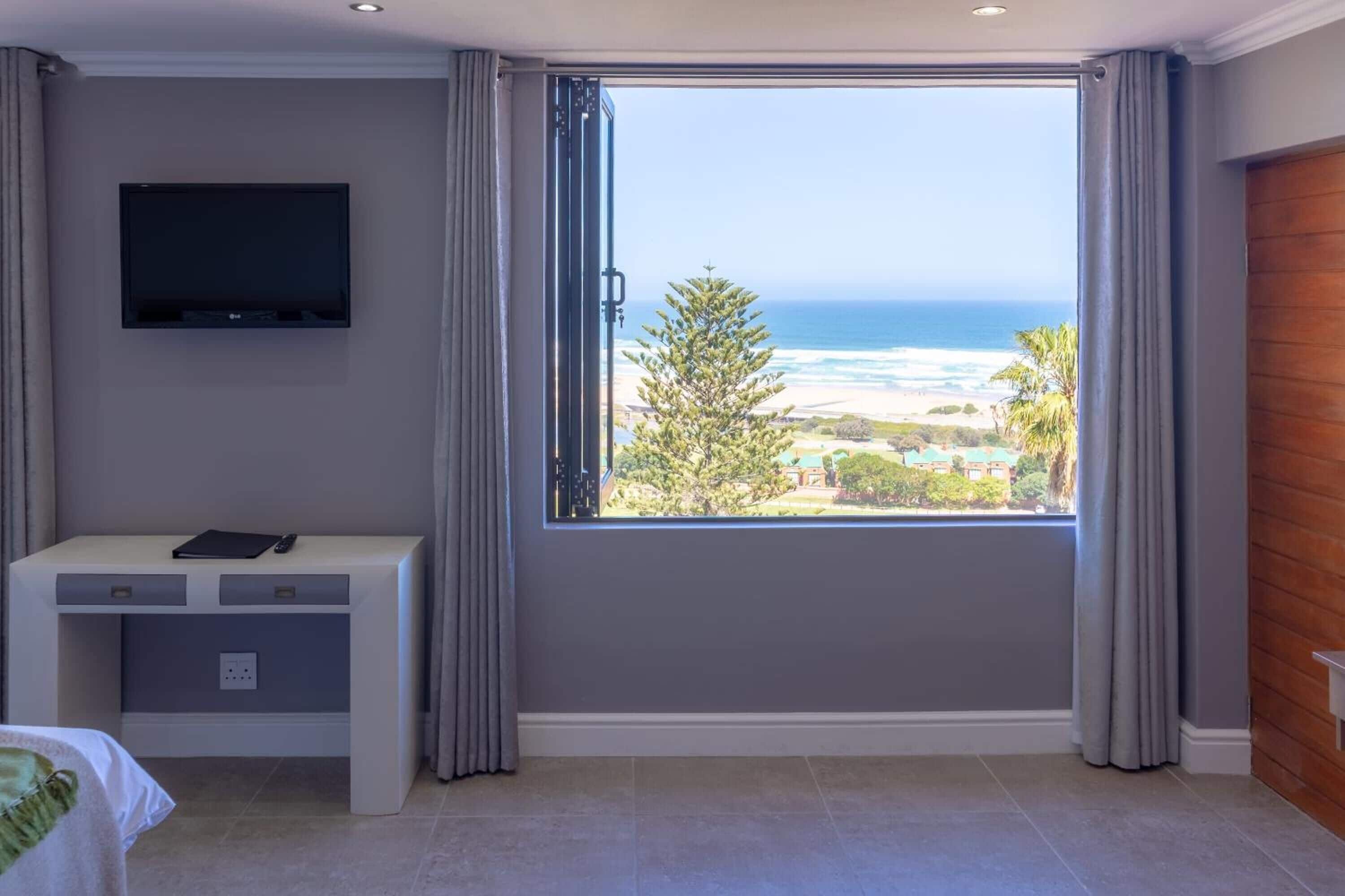 Quarto com uma grande janela com vista para o oceano e a praia, uma televisão de tela plana na parede e uma mesa de cabeceira com abajur.