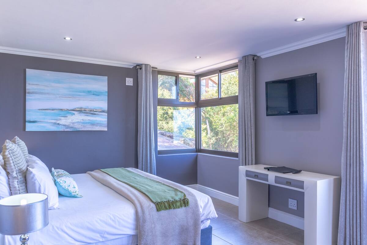 A bedroom with a bed, a desk, a TV, and a window with curtains.
