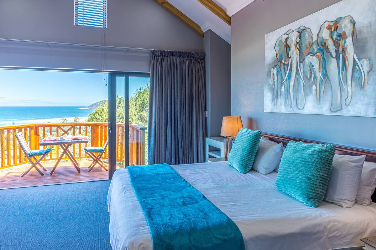 A bedroom with a large bed, a view of the ocean, a balcony with chairs, and a painting on the wall.