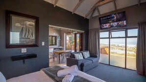 A hotel room with a bed, a gray sofa, a dining area, and a large window with a view. - Boardwalk Lodge - B&B (Wilderness)