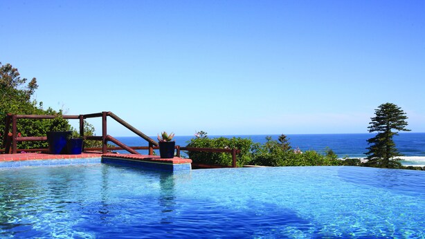 A infinity pool with a wooden deck and potted plants, overlooking a beach and ocean. - Boardwalk Lodge - B&B (Wilderness)