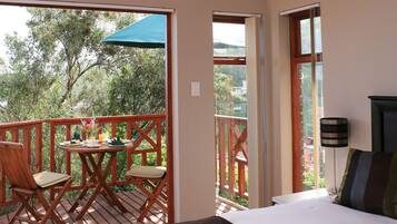 A bedroom with a large bed, a balcony with wooden furniture, and a view of trees.