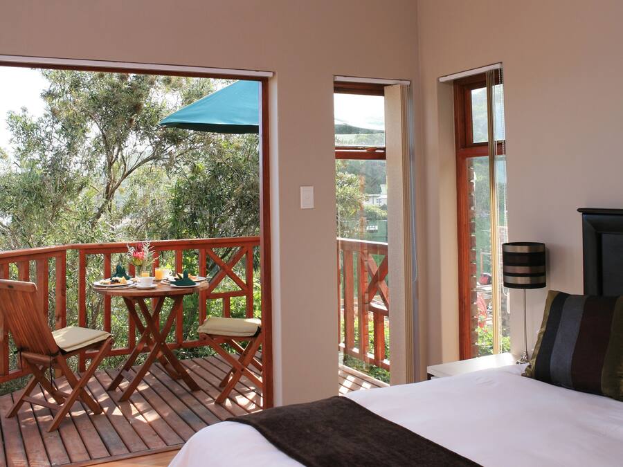 A bedroom with a large bed, a balcony with wooden furniture, and a view of trees.
