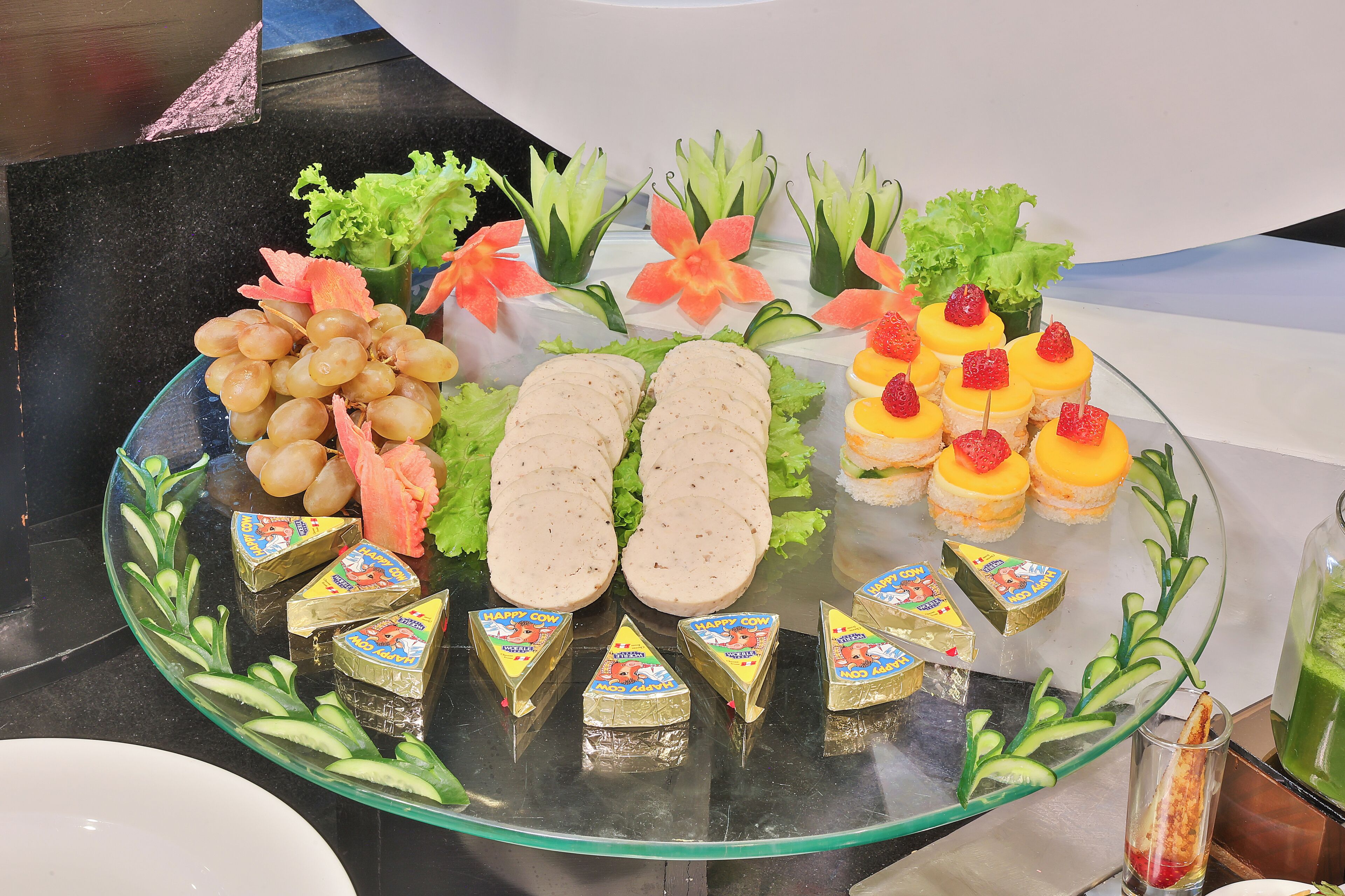 A glass platter with various food items, including sliced meats, fruits, and decorative flowers.