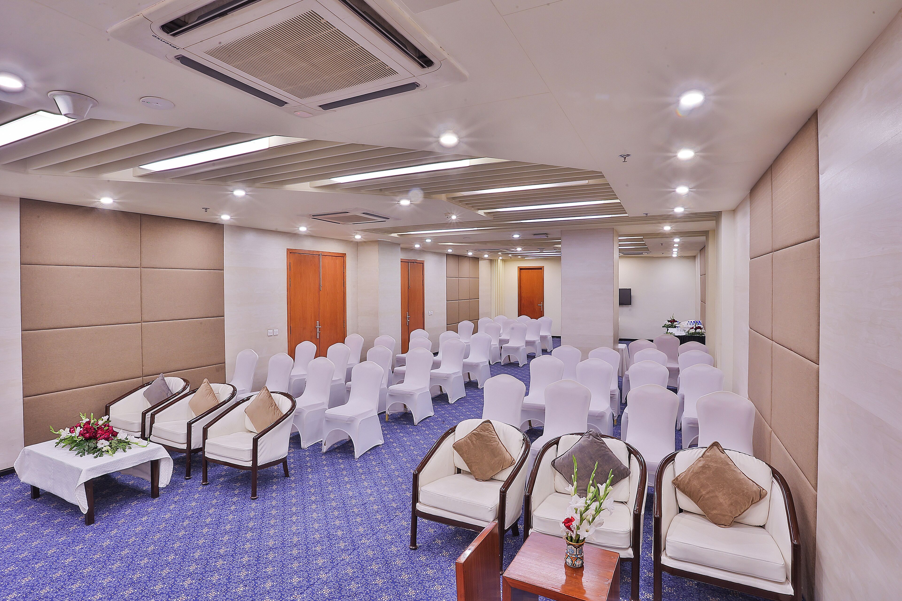 A conference room with rows of white chairs, a few sofas with cushions, a small table with a flower arrangement, and a carpeted floor.