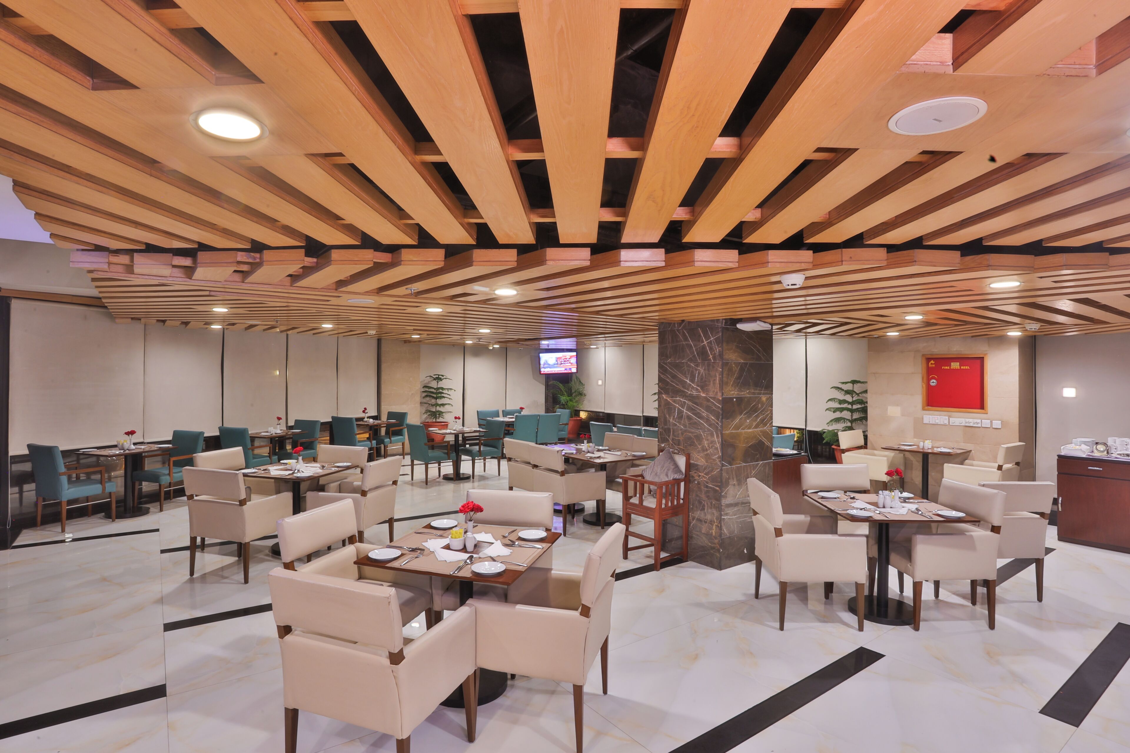 A modern restaurant with wooden ceiling beams, multiple dining tables with chairs, and a TV screen in the background.