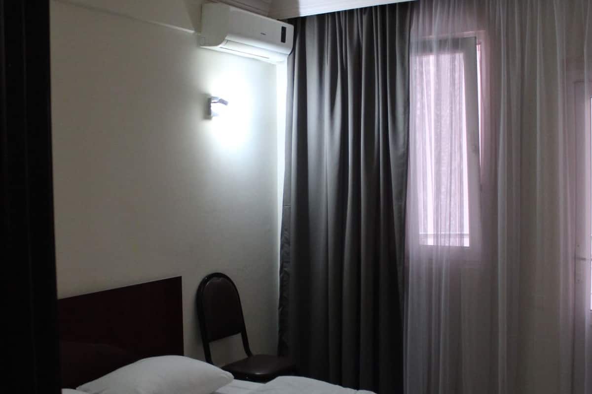 A hotel room with a bed, a chair, a window with curtains, and an air conditioning unit.