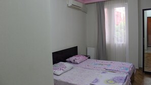 Quarto com duas camas, janela com cortinas, aparelho de ar condicionado e porta para outro cômodo.