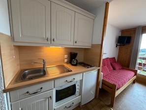 A compact kitchenette with a sink, stove, and oven, a bed with red polka dot bedding, and a TV mounted on the wall.