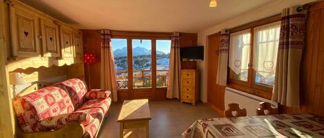 A cozy room with a wooden sofa, a small table, a TV, and a window with a mountain view.
