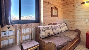 A cozy cabin room with a wooden interior, a bed with patterned pillows, a small wooden chair, and a window offering a mountain view. - Family apartment near slopes (La Plagne)