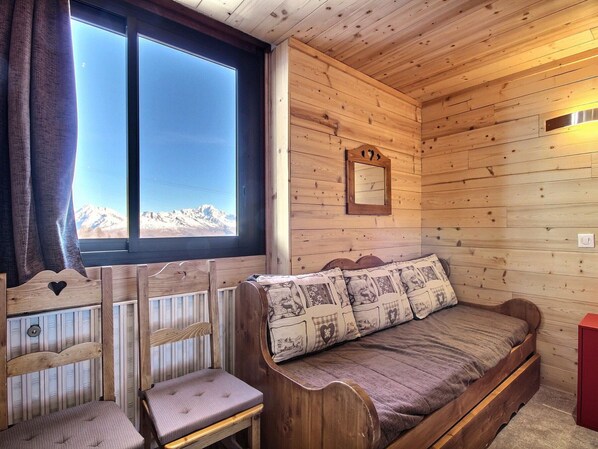 A cozy cabin room with a wooden interior, a bed with patterned pillows, a small wooden chair, and a window offering a mountain view.