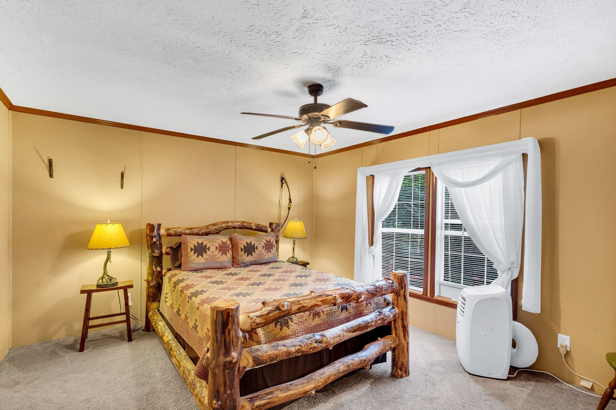 A bedroom with a rustic wooden bed, a ceiling fan, a window with white curtains, and a bedside lamp.