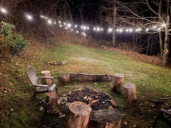 A nighttime outdoor setting with a fire pit surrounded by wooden stumps, a chair, and string lights. - Our secluded Cabin in Maggie Valley, NC (maggie valley)