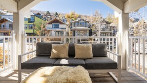 A wooden deck with a gray sofa and fluffy white rug, overlooking a row of colorful houses. - Walk to Trails & Main St, 4BR Hot Tub Home (Park City)