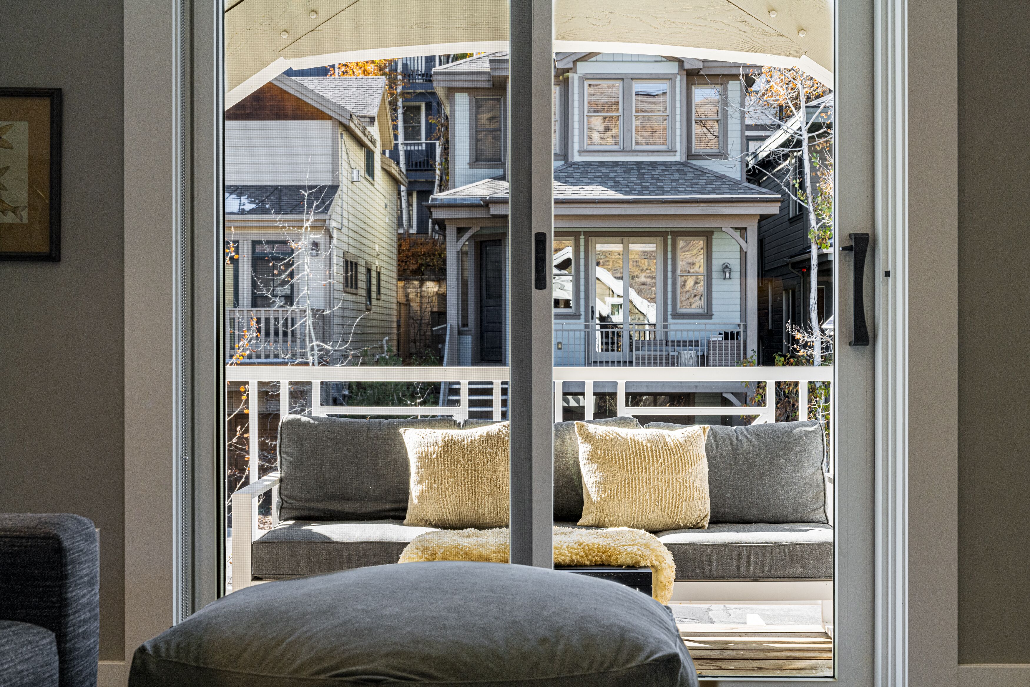 A view through a glass door to a balcony with a sofa and cushions, overlooking a residential area with houses and a tree.