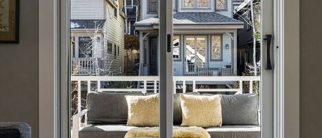 A view through a glass door to a balcony with a sofa and cushions, overlooking a residential area with houses and a tree.