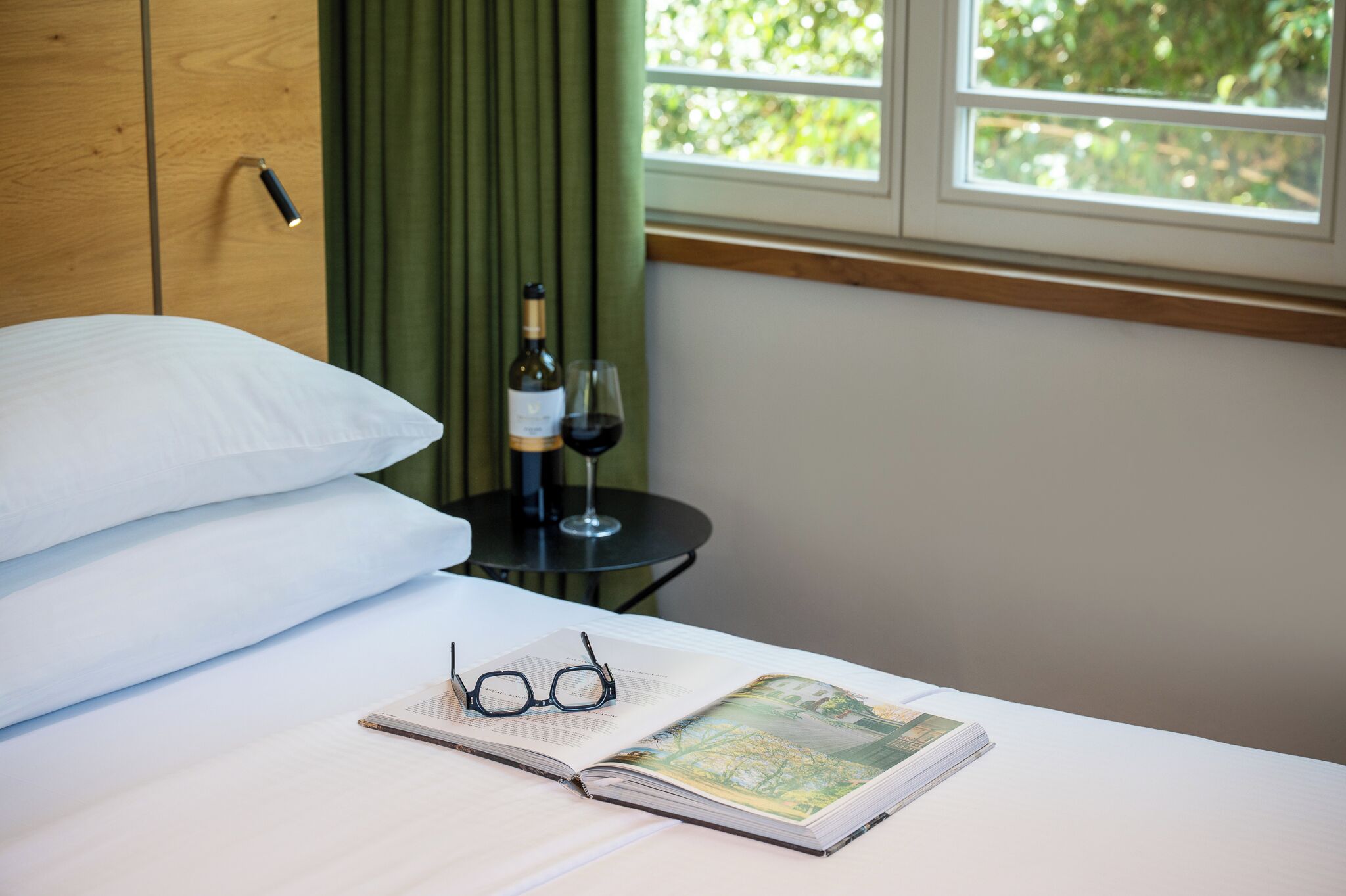 A neatly made bed with pillows, a book, glasses, a wine bottle, and a glass on a bedside table.
