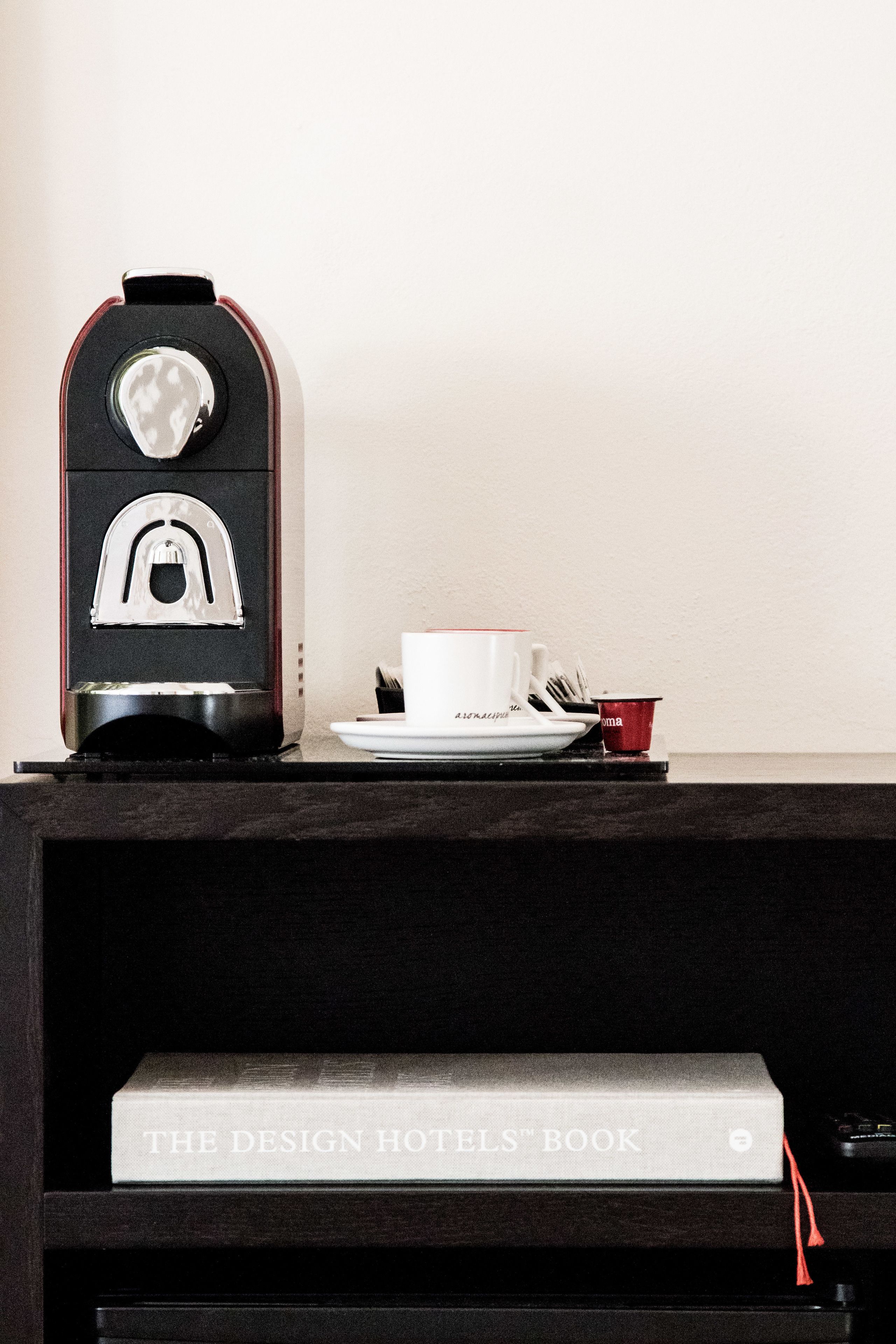 Une machine à café, une tasse blanche, une tasse rouge et un livre intitulé « The Design Hotels Book » sont posés sur une étagère.