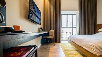A hotel room with a bed, a desk with a laptop, a chair, a TV mounted on the wall, and a window with curtains.