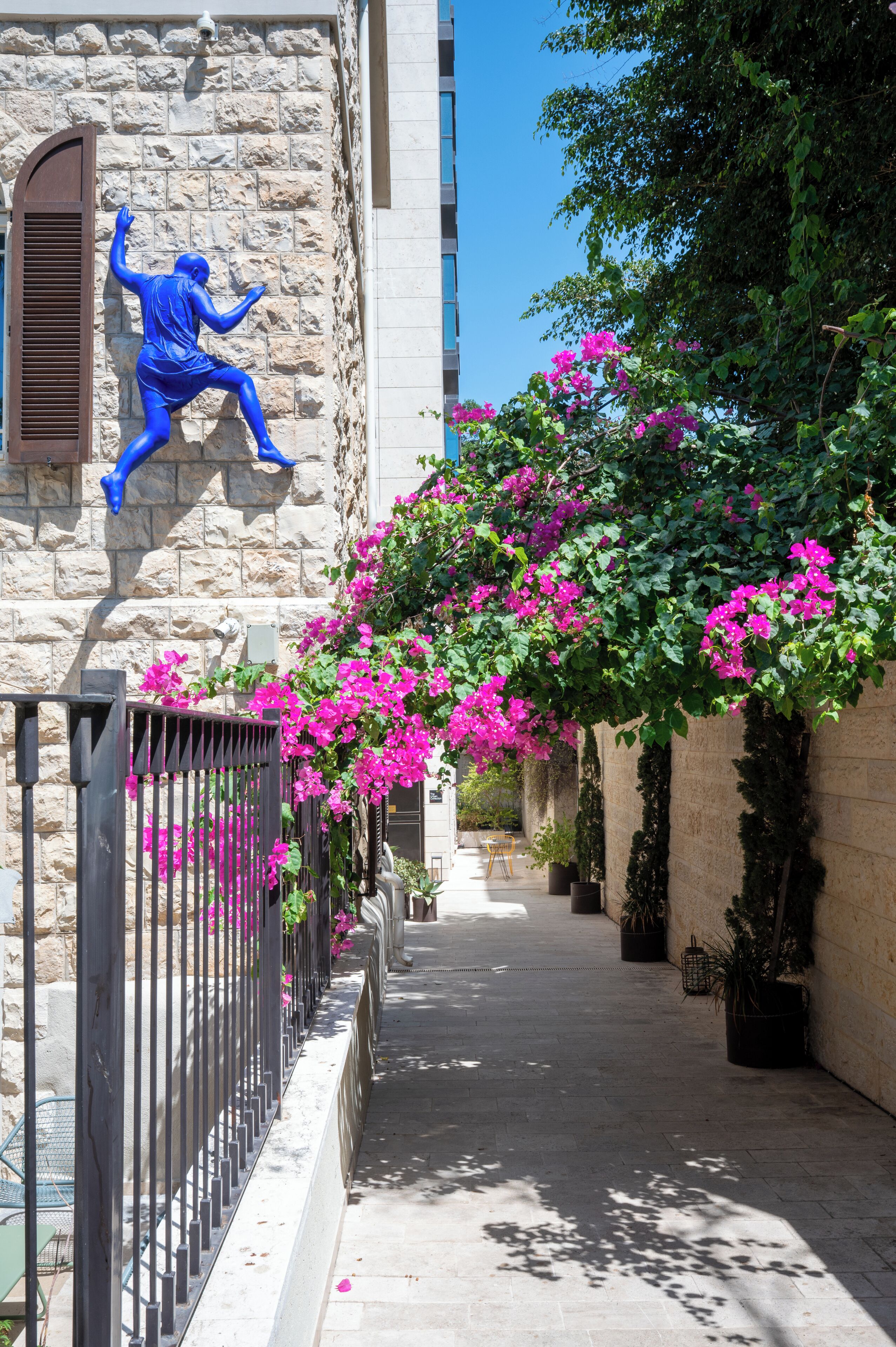 A narrow alleyway with a stone building, a blue statue, and vibrant pink flowers.
