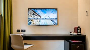 A hotel room with a flat-screen TV mounted on the wall displaying a cityscape, a desk with a chair, a coffee machine, and a book titled "Motel Restaurant Bar Club Design".