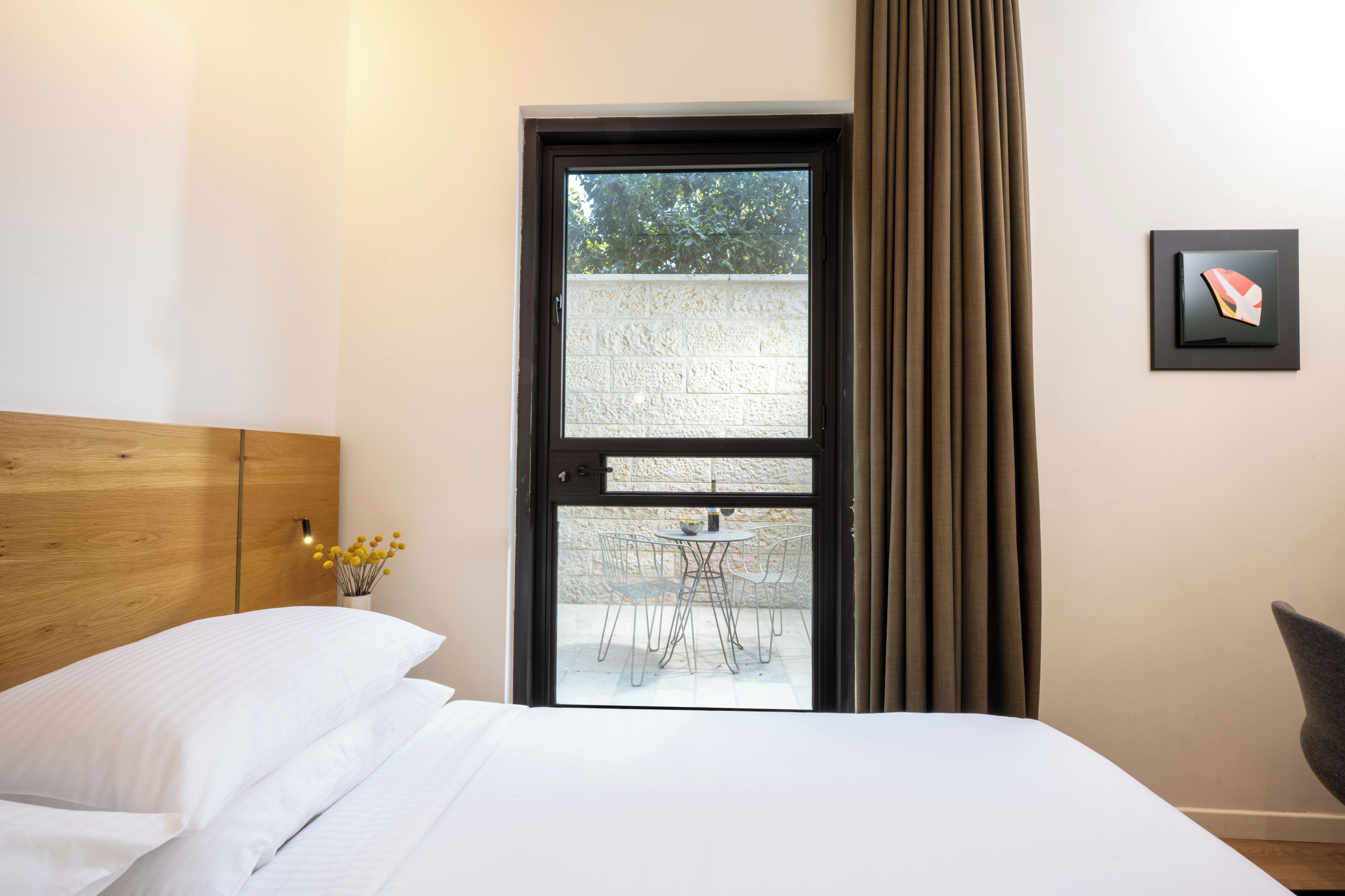 Habitación de hotel con cama, cabecera de madera, ventana con cortinas, vista a un área de estar al aire libre y un cuadro en la pared.
