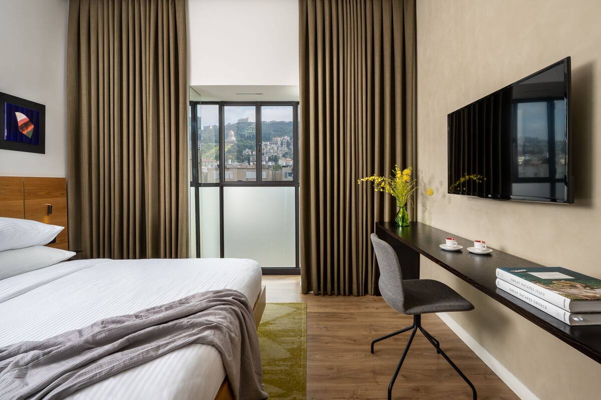 A hotel room with a bed, a desk with a chair, a TV, and a window with curtains.