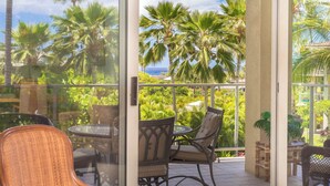 A balcony with a glass door, furnished with wicker chairs and a table, overlooking a lush garden and palm trees. - Charming Renovated Condo at Vista Waikoloa! (Waikoloa)