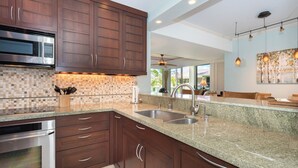 A modern kitchen with dark wood cabinets, a granite countertop, stainless steel appliances, and a large window offering a view of the outdoors. - Charming Renovated Condo at Vista Waikoloa! (Waikoloa)