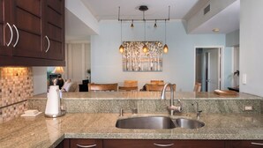 A modern kitchen with granite countertops, stainless steel sink, and wooden cabinets. There is a dining area with a sofa and table in the background. - Charming Renovated Condo at Vista Waikoloa! (Waikoloa)