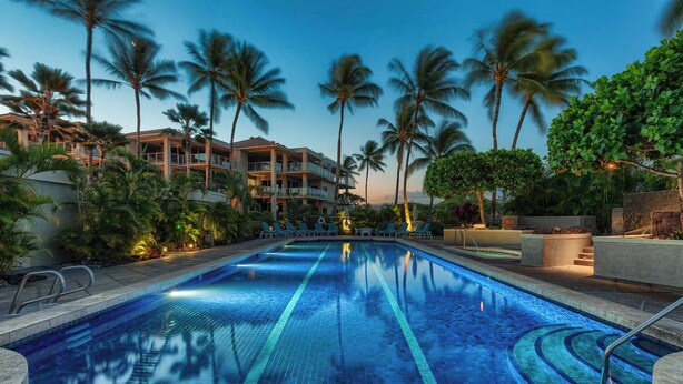 A swimming pool with clear blue tiles, surrounded by palm trees and a multi-story building with balconies. - Charming Renovated Condo at Vista Waikoloa! (Waikoloa)