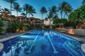 Una piscina con baldosas azules brillantes, rodeada de palmeras y un edificio de varios pisos con balcones.
