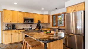 A modern kitchen with wooden cabinets, a granite countertop, a stainless steel refrigerator, and a stone floor. - RMR: Three Bear Tracks Home in Teton Village (Teton Village)