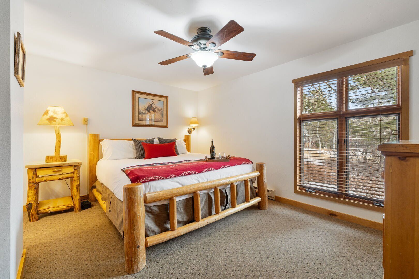 A bedroom with a wooden bed frame, a ceiling fan, a bedside table with a lamp, a window with blinds, and a framed picture on the wall.