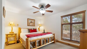 A bedroom with a wooden bed frame, a ceiling fan, a bedside table with a lamp, a window with blinds, and a framed picture on the wall. - RMR: Three Bear Tracks Home in Teton Village (Teton Village)