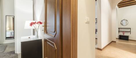 Un pasillo con puerta de madera, pared blanca, espejo y una pequeña mesa con un jarrón de flores.