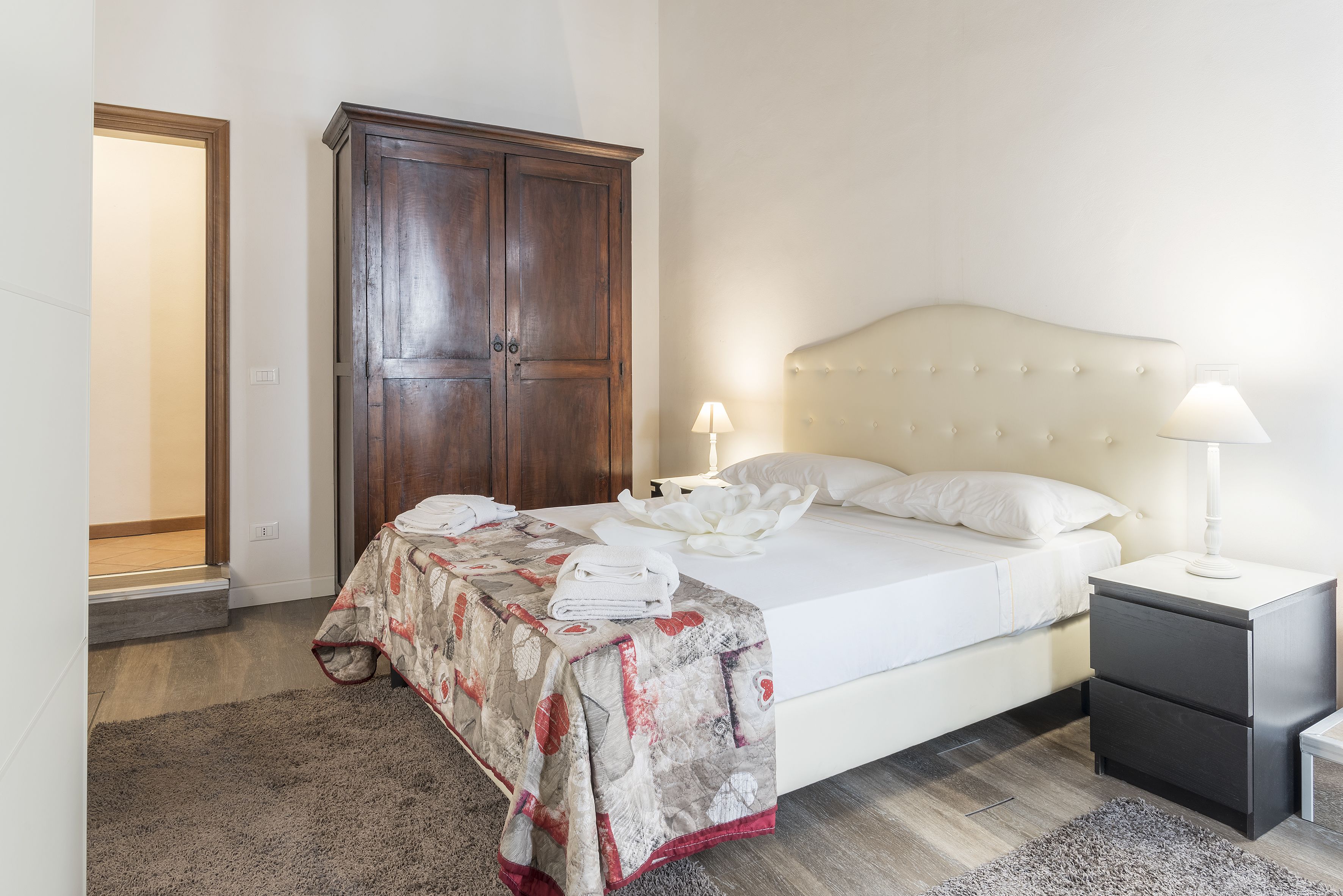 A neatly made bed with a white comforter and pillows, a bedside table with a lamp, a dark wooden wardrobe, and a grey carpet.